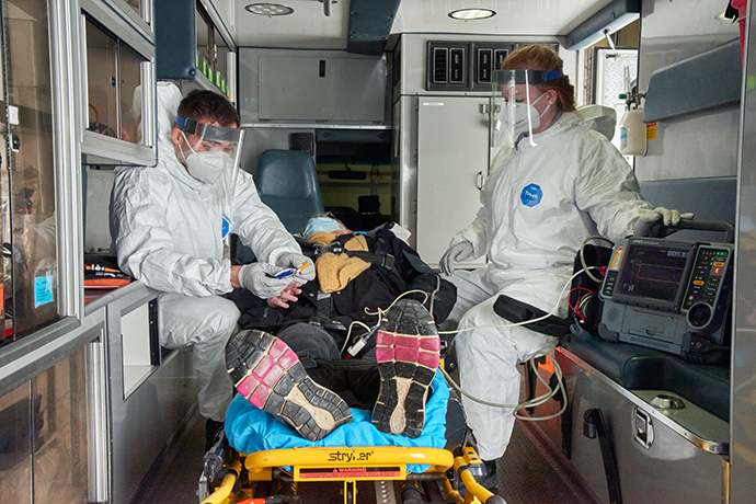 First responders in protective gear, face masks and shields taking a peron&#039;s vitals in the back of an ambulance