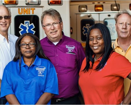Diverse Universal Ambulance Services employees in different colored shirts smiling at the camera in front of some ambulances